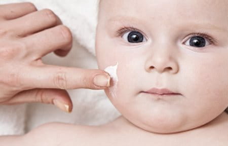 La mano de un adulto aplicándole crema al rostro de un bebé.